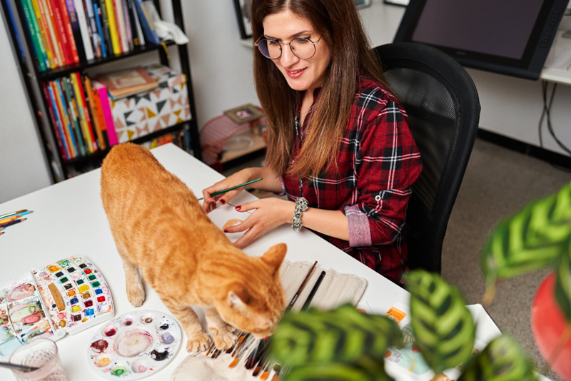 Artist stops her work because her big orange cat walks across the table, portrayed by a cat model and a woman model.