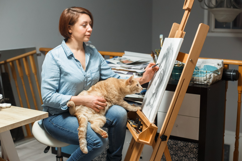Woman sitting and drawing on a canvas, her cat on her lap is smelling the canvas way too close, illustrated by two models.
