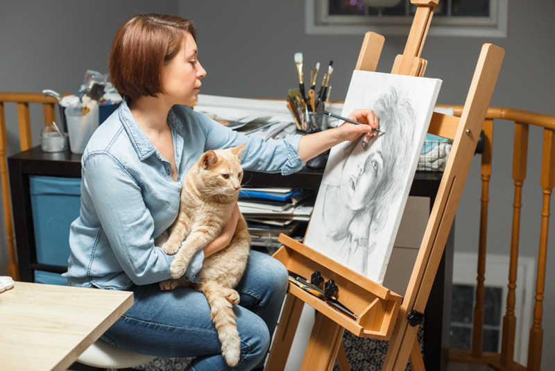 Artist is drawing on a canvas while her orange cat on her lap looks at it with intent, embodied by a cat model and a woman model.