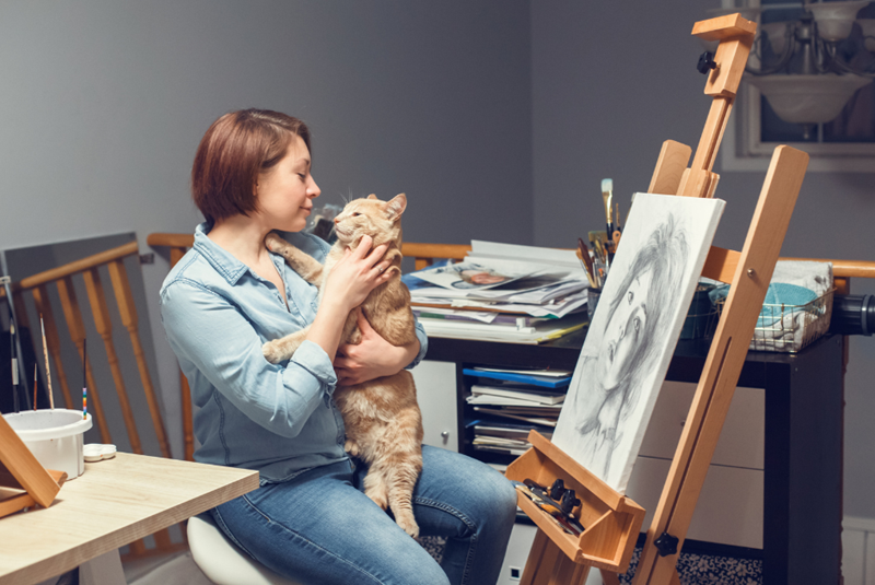A woman stops drawing in the middle of the process because her cat jumped on her lap and demanded pets, personified by a couple of feline and human models.