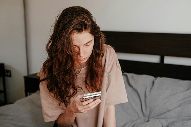 Woman sitting on a bed and looking down at her phone with a serious expression.