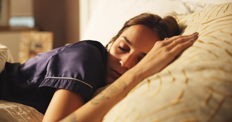 Woman in dark pajamas sleeping on a bed, resting her head on a pillow in warm indoor light.