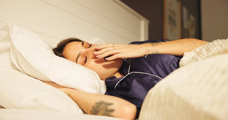 Woman in dark pajamas yawning in bed under white covers with warm light in a hotel room.