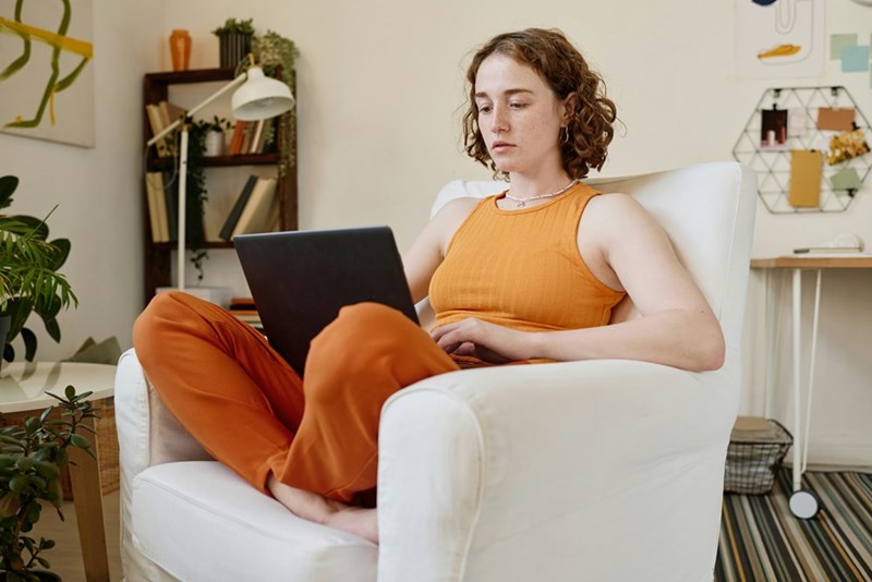 Woman sitting in a white armchair at home and working on a laptop.