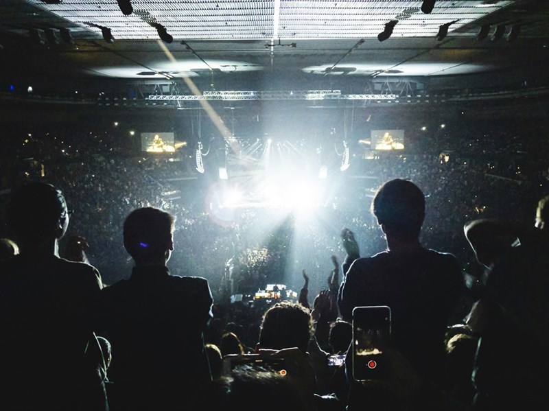 Crowd watching a concert in a packed arena with bright stage lights and people filming on phones.