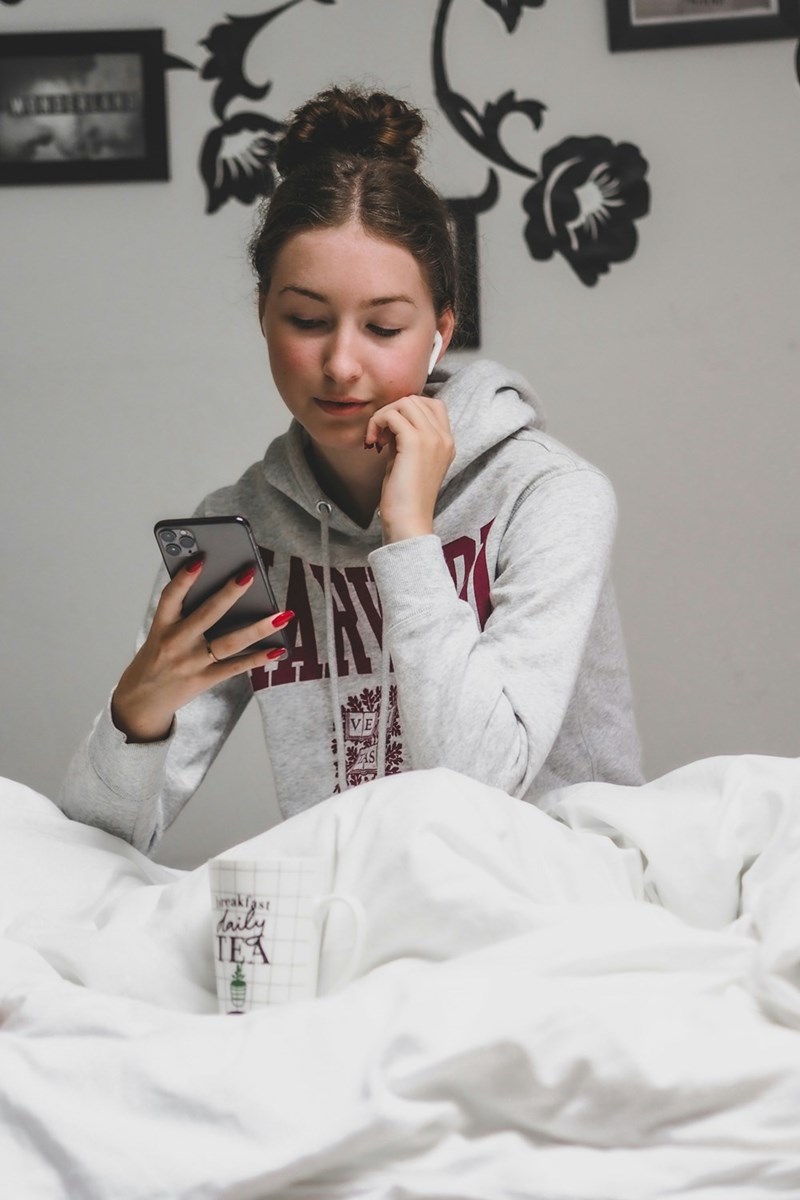Young woman sitting in bed with a phone, wearing a gray hoodie and earbuds.
