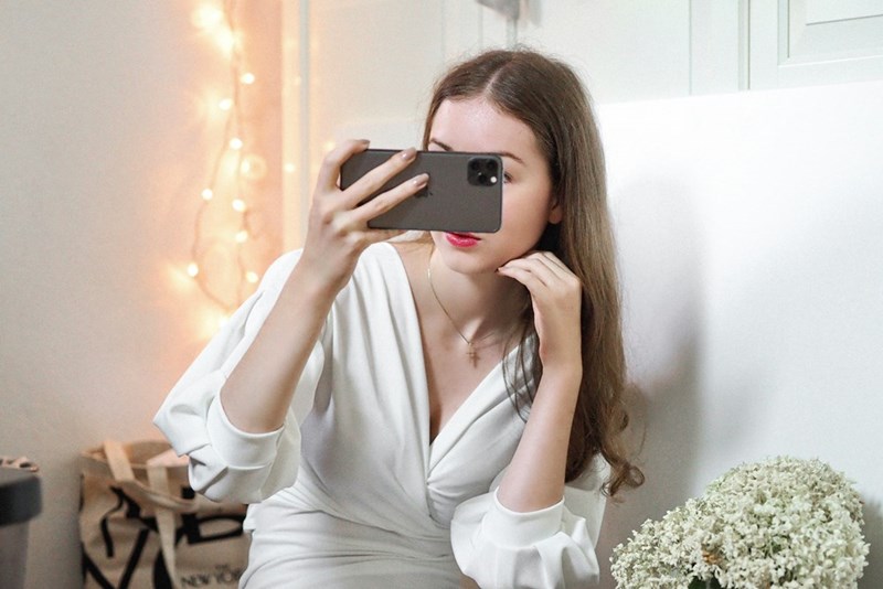Woman in a white top holding up her phone while sitting indoors near string lights.
