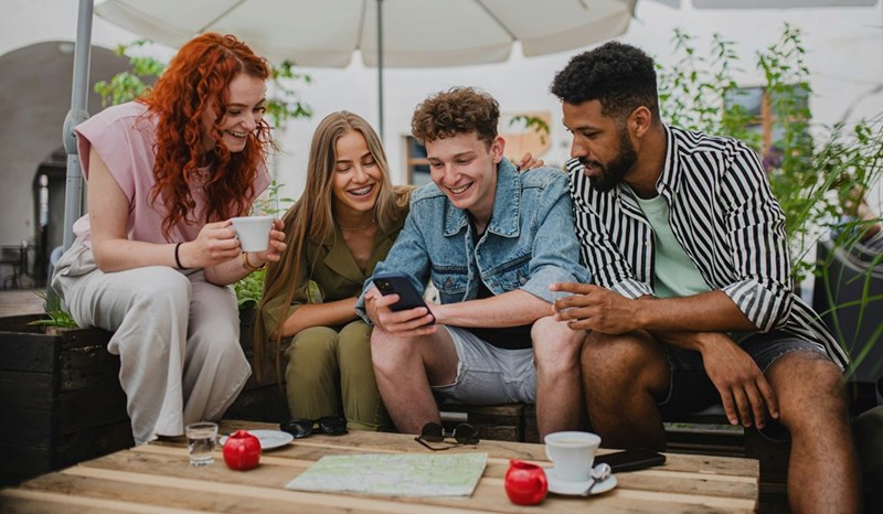 A group of friends sitting down at a restraunt table smiling and looking at a phone one of them holds.