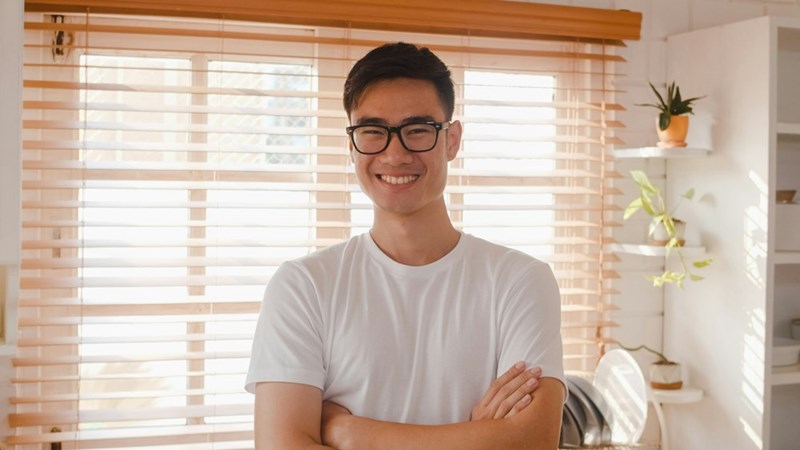 The portrait shows a young professional in a bright home setting, reflecting the optimism often seen at the start of a long-distance relationship.