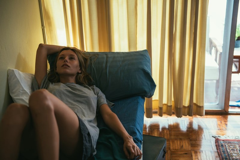 Woman lying on a couch indoors with one arm behind her head, looking upset or deep in thought.