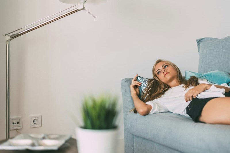 Woman lying on a light blue couch and looking thoughtful in a bright living room.