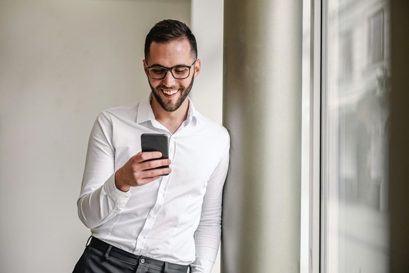 Smiling man in glasses and a white shirt looking at his phone while standing by a window.