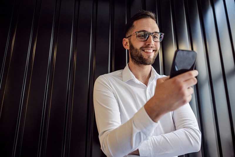 Man in a white shirt and glasses looking at his phone while standing indoors against a dark paneled wall, like in a hotel lobby maybe
