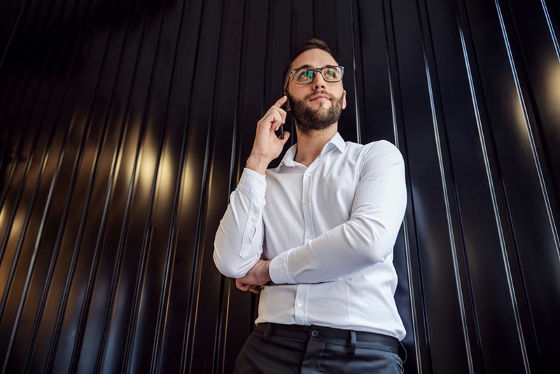 Man in a white shirt and glasses talking on the phone against a dark paneled wall.