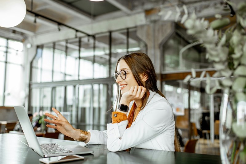 Woman in glasses smiling at a laptop in a bright office, resting her chin on her hand.