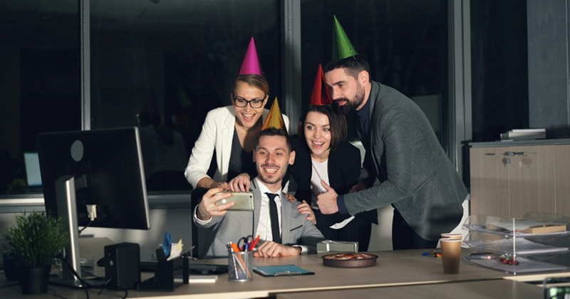 Models represent four colleagues gathering around a desk at night, taking a selfie while wearing party hats