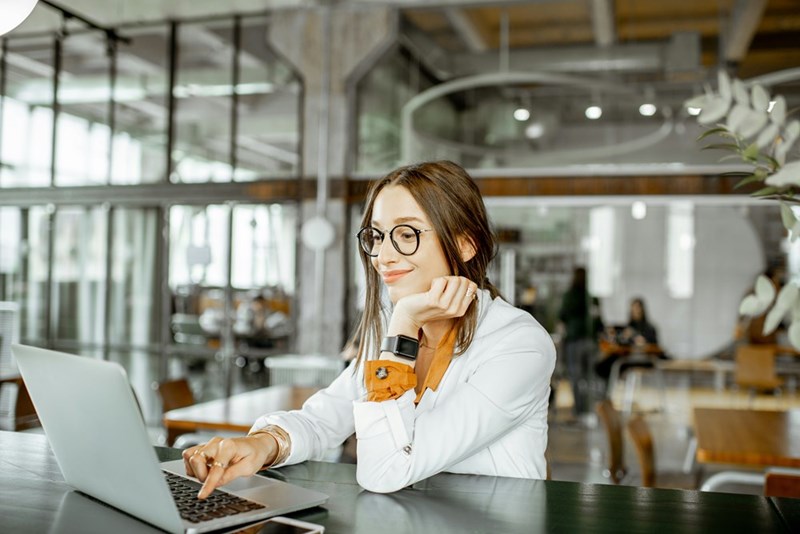 Woman in glasses smiling at a laptop in a modern office, resting her chin on her hand.