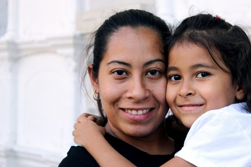 Woman and her toddler daughter smiling hugging each other while looking at the camera.