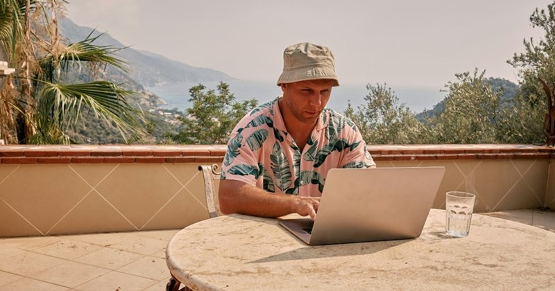A man sits at a table with his laptop computer while on vacation. The subject is a model.