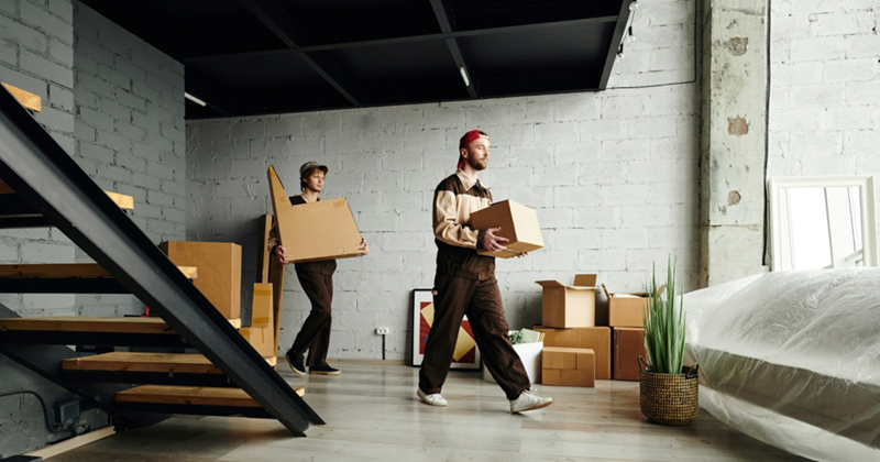 A representation of two men carrying boxes and furniture into a large storage room with a staircase