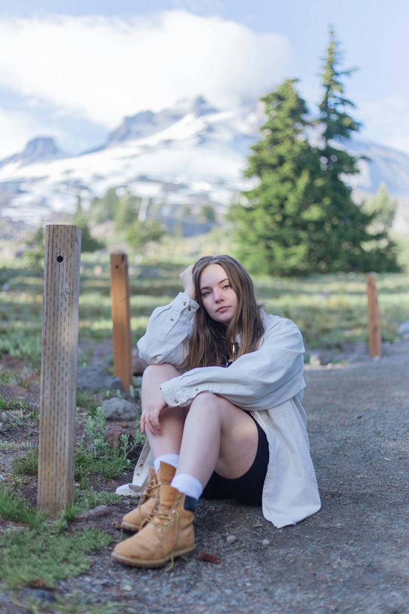 Woman sitting alone outdoors with her hand on her head and snowy mountains blurred behind her.