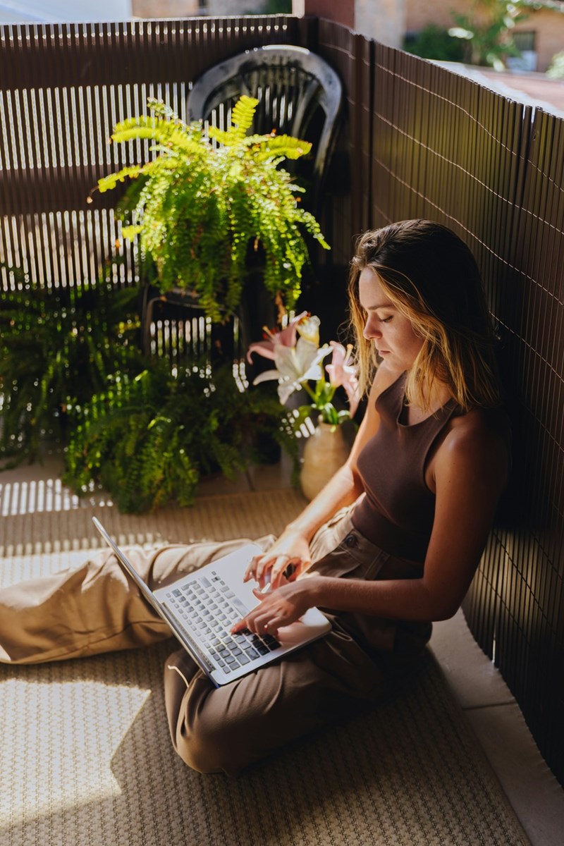 Woman sitting on a balcony with a laptop, surrounded by plants in warm sunlight.