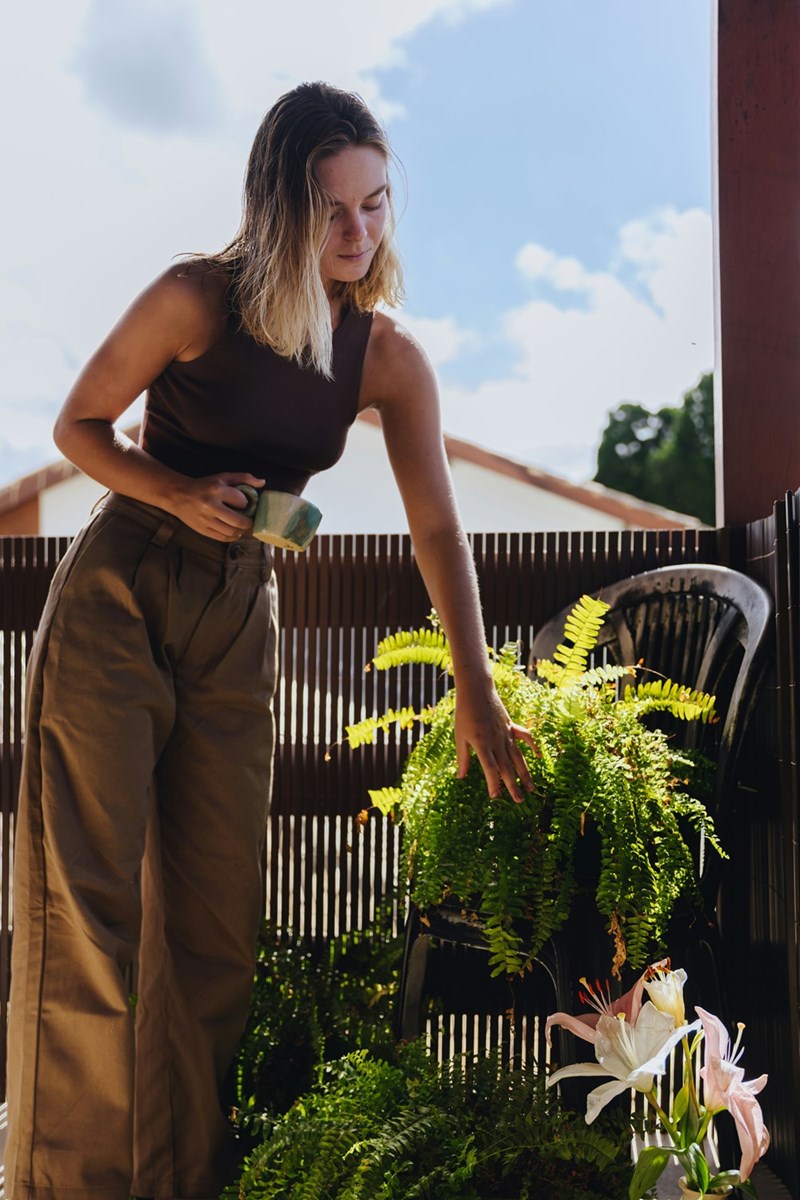 Woman standing on a balcony with a mug, touching green plants in bright sunlight.