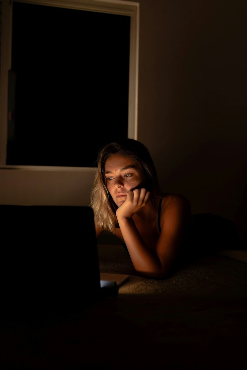 Woman lying in bed at night and looking at a laptop, lit by the screen in a dark room.