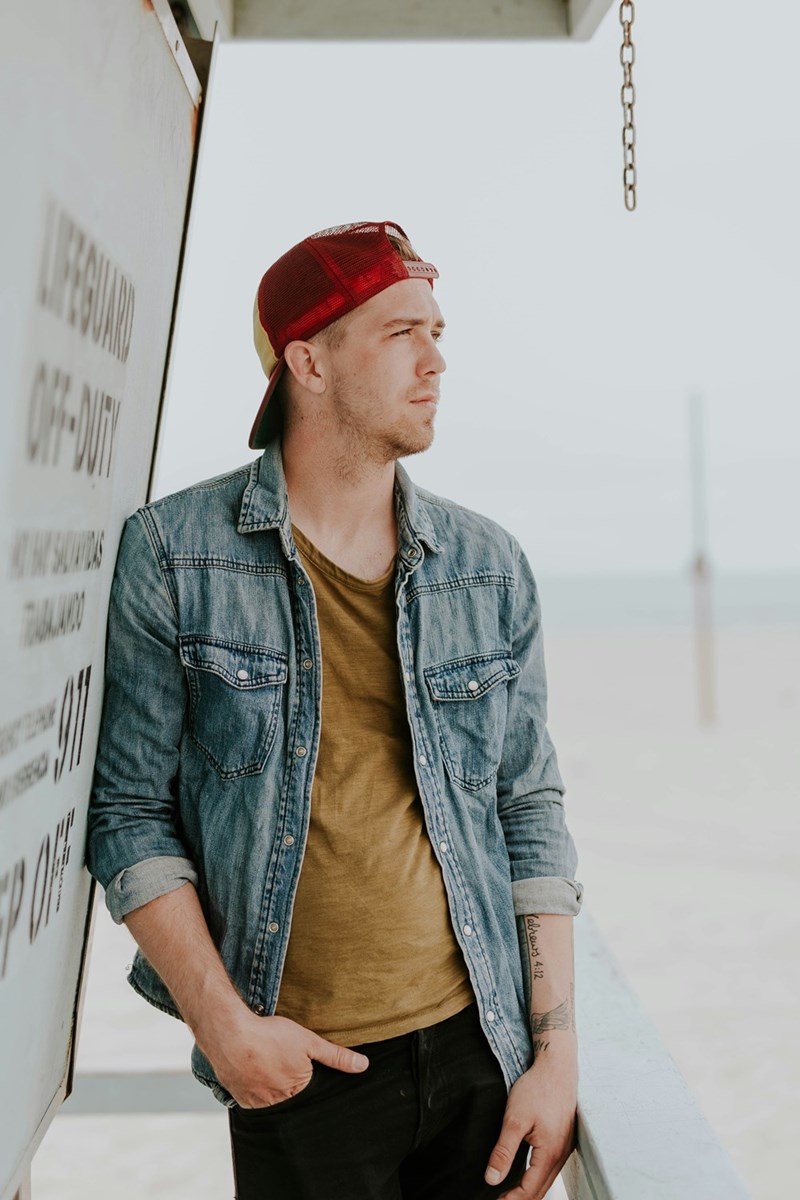 Man in a denim shirt and red cap standing near a beach with a serious expression.