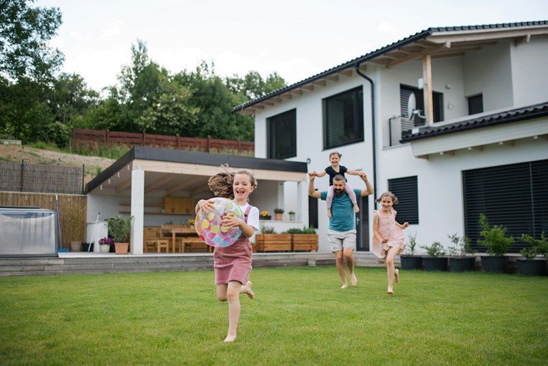 Kids and father play with a beach ball outside of their spacious home and grassy back yard, as demonstrated by models.