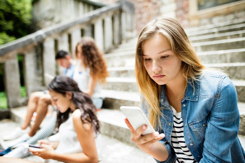 University student is engrossed in her phone while sitting outside with friends, as a model depicts.