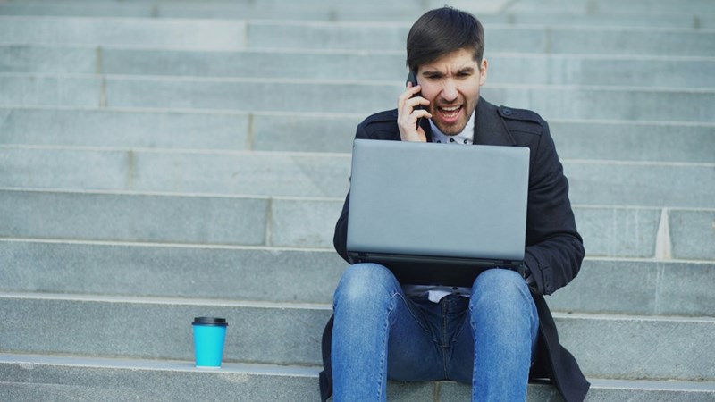 A photograph representing an angry employee sitting on the steps of a building screaming on his phone while working on his laptop.