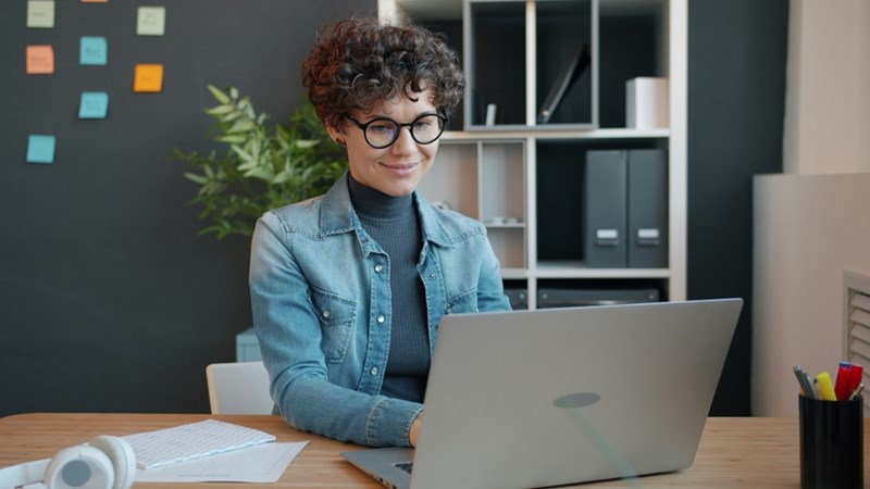 Woman with short curly hair wearing glasses sitting at an office looking at the computer.