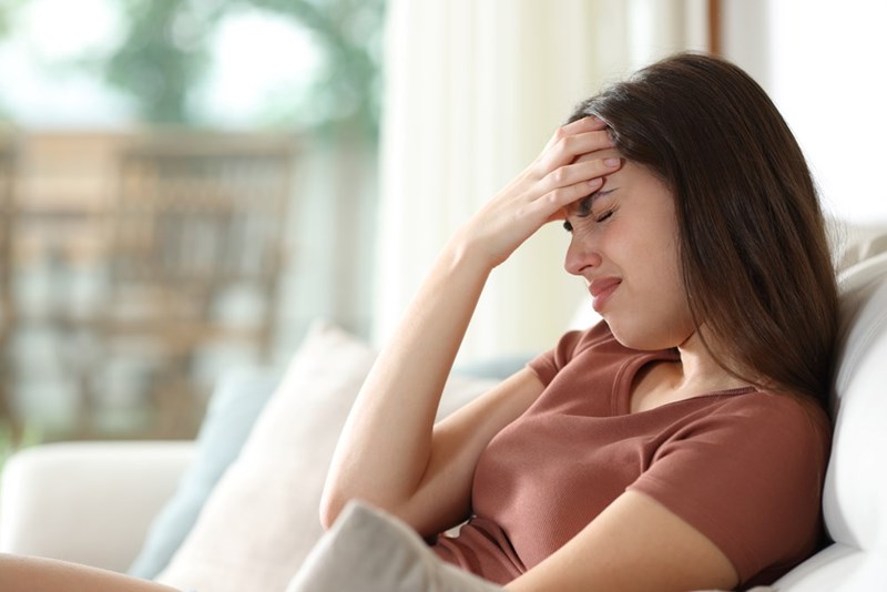 Woman sitting on a couch with her hand on her forehead, looking upset in a bright living room.