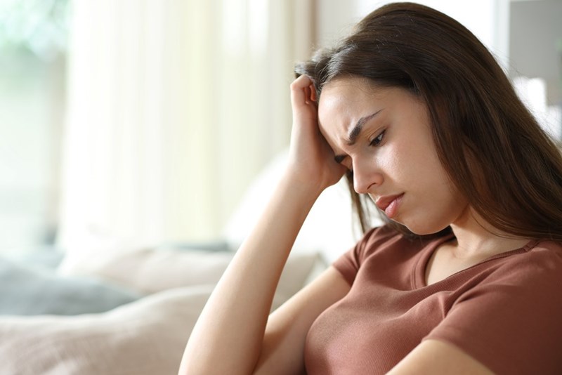 Woman sitting on a couch with her hand in her hair, looking upset in a bright living room.