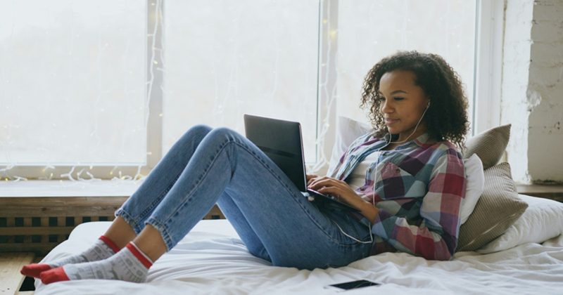 A representation of a young woman reclining on a bed, propped up by cushions, and using the laptop on her lap