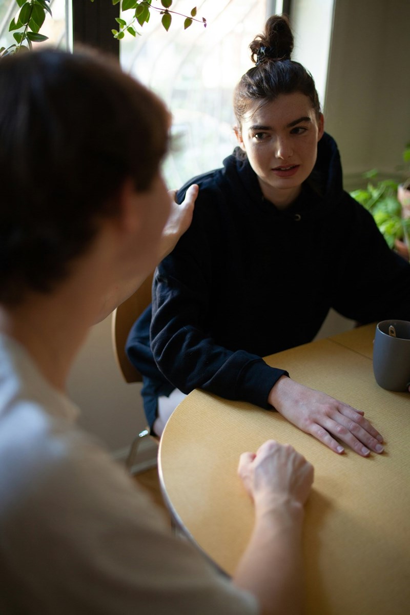 Woman in a black hoodie sitting at a table during a serious conversation indoors.