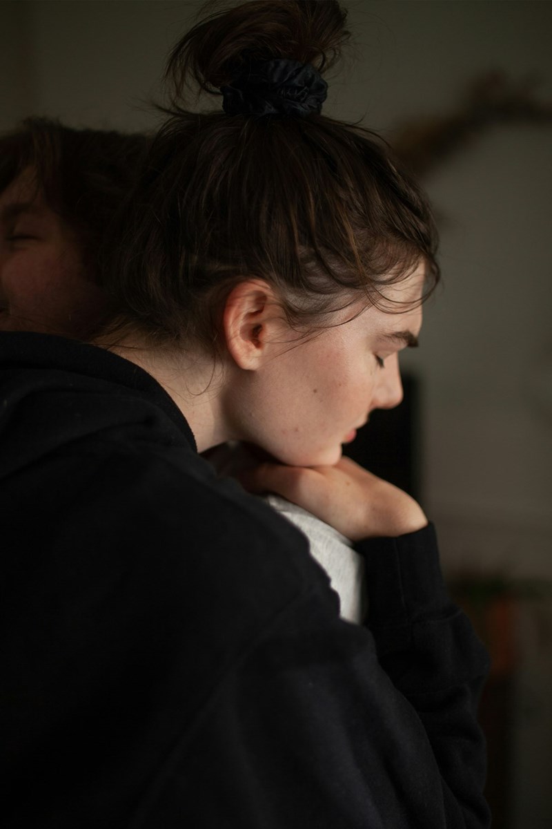 Woman in a black hoodie being comforted with a hug, looking down sadly indoors.