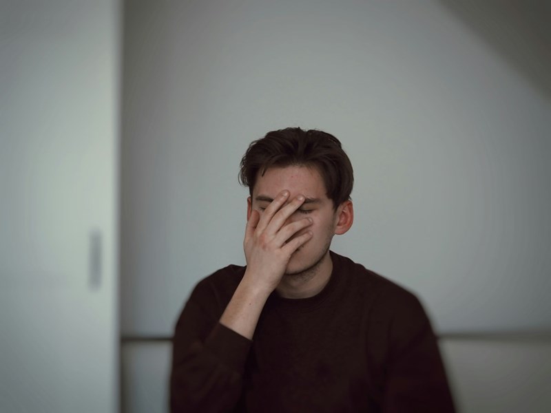 Man sitting indoors with his hand covering his face, appearing stressed and overwhelmed.