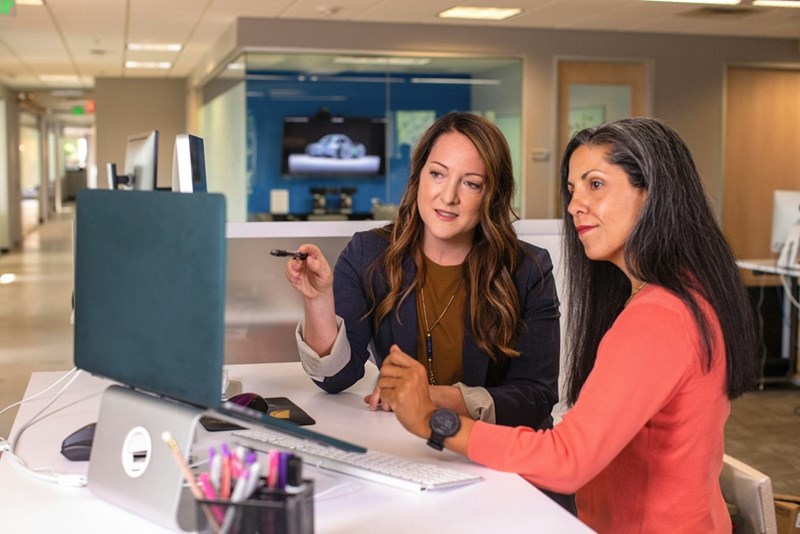 Two coworkers review information together on a desktop computer during a discussion at the office.