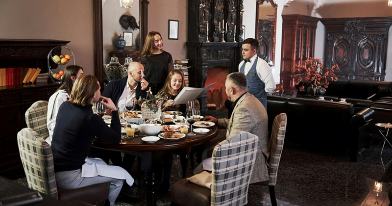 A representation of a group of people sitting at a round table, having their order taken by a server in a restaurant