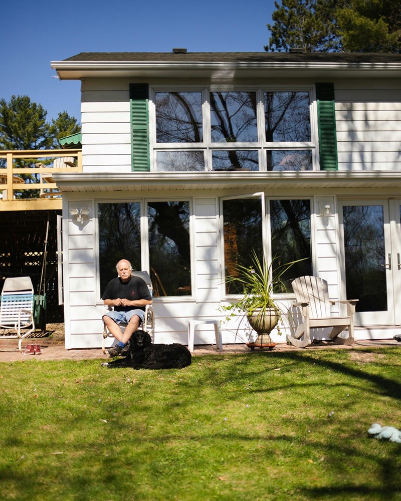 A photograph of an older man sitting in a chair outside his house overlooking his lawn.