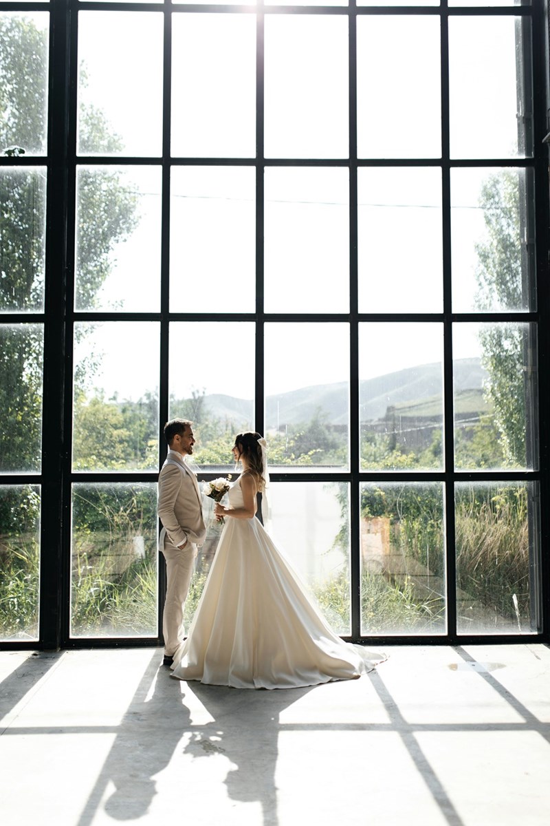 Couple pose for wedding photos in front of a large glass window, as depicted by models.
