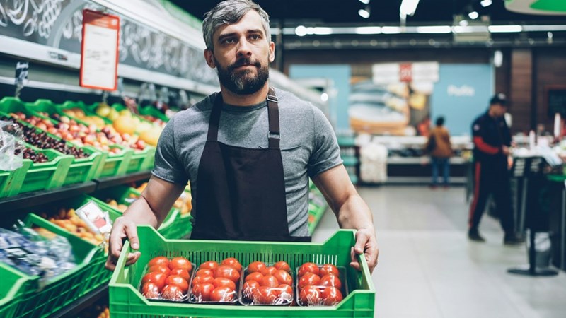Grocery store worker stocks produce while looking unhappy about his work situation, in a representation by a model.