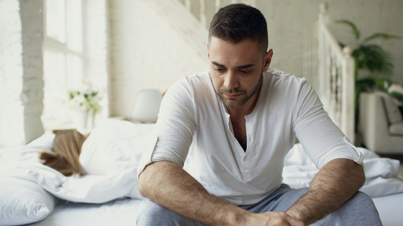 Man wearing white shirt and joggings sitting on the couch upset looking down.
