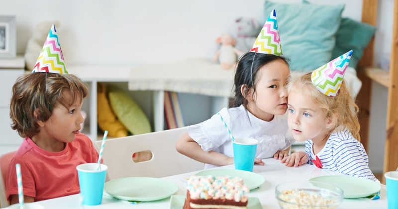 A representation of three young girls sitting at a table wearing party hats, with two of the girls whispering to one another