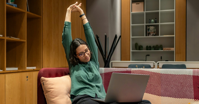 A model representing a young female employee sitting on a couch with a laptop on her lap while stretching