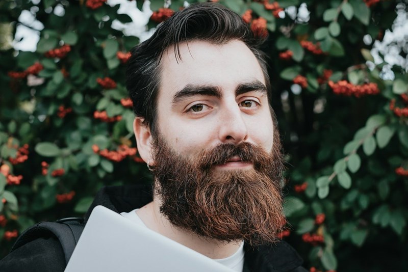A close-up portrait captures a bearded man standing in front of lush greenery and bright red berries.