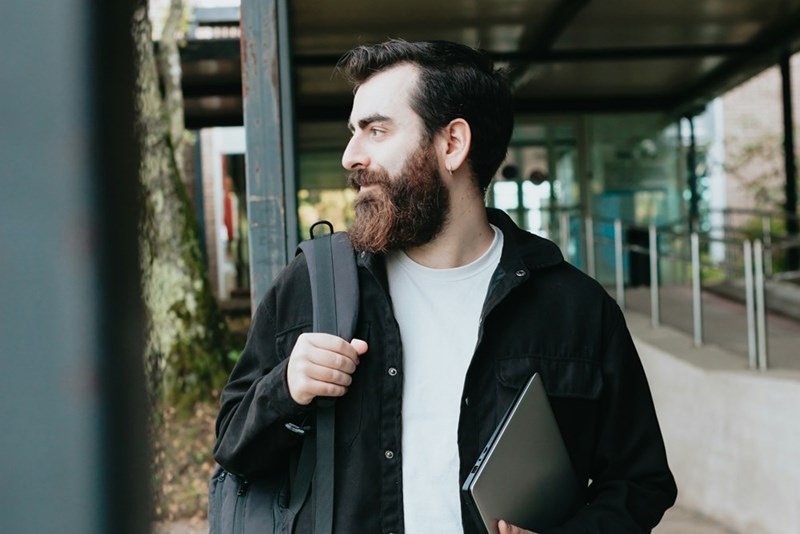 A man carrying a backpack and laptop walks through an urban area, appearing ready for work or study.