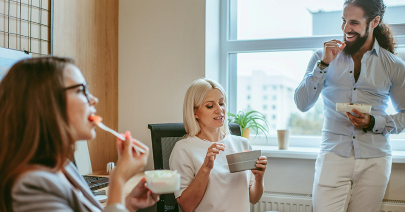 A representation of two female coworkers sitting down and eating from lunch boxes, with a male coworker standing beside them while also eating from a lunchbox